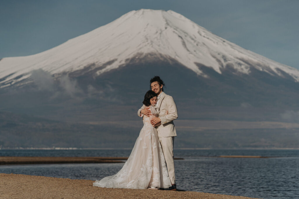 A couple in white wedding dress and cream suit embrace under a snow capped Mt. Fuji during their Japan Elopmenet