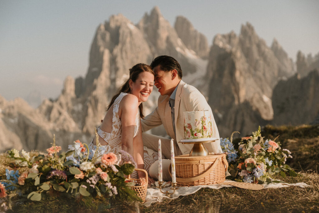 A couple sit down to a picnic with cake and flowers on their wedding day with the mountain peaks of Cadini di Misurina behind them