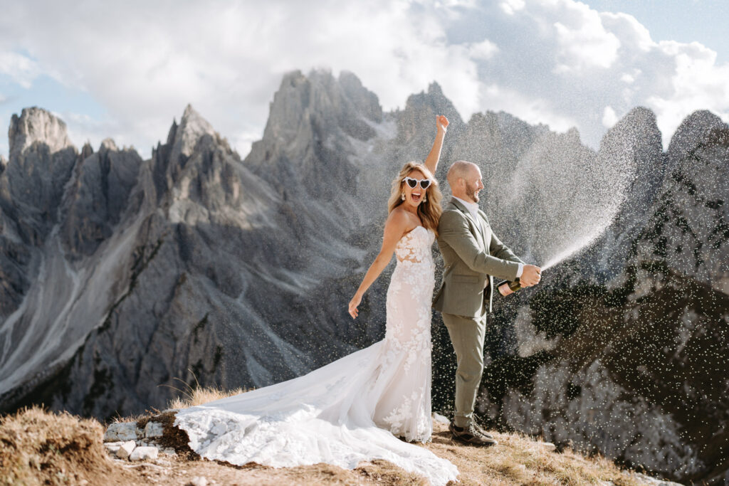 A couple eloping in the Dolomites spray champagne after their vows with the jagged mountain peaks of Cadini di Misurina behind them 