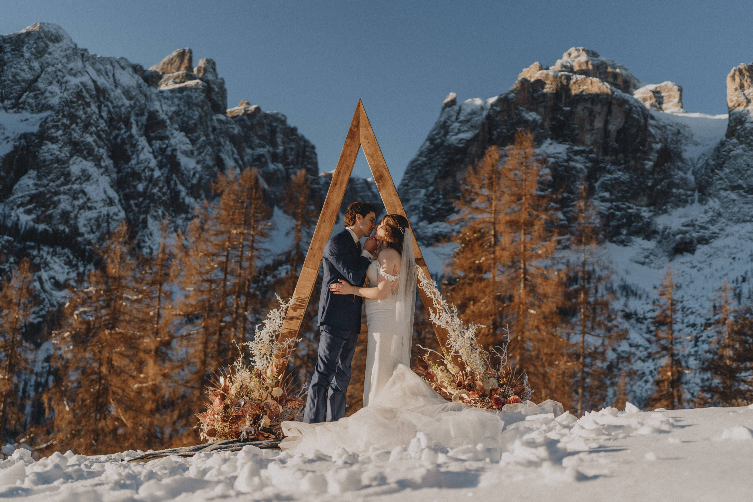A couple kiss under a wooden arch, framed by flowers and snow on a clear winter morning in the Dolomites