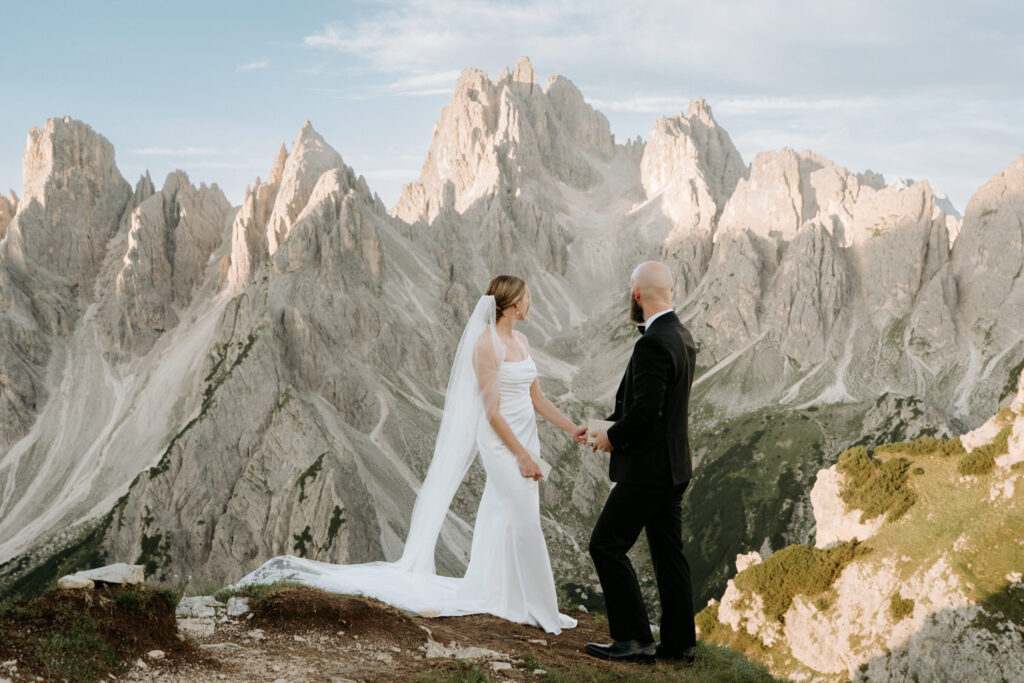 A wedding couple wearing white dress and black suit stand on a cliff edge looking off over sunlit mountain peaks in the Italian Dolomites
