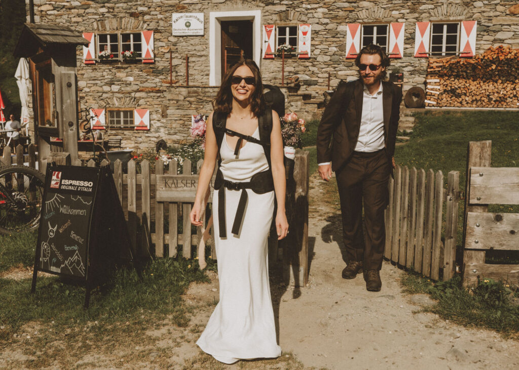 A couple in wedding dress and brown suit walk out of a traditional Austrian mountain hut on their elopement day, smiling