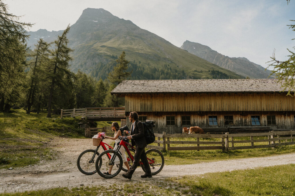 A couple walks two red mountain bikes down a gravel path in Austria on their elopement day.