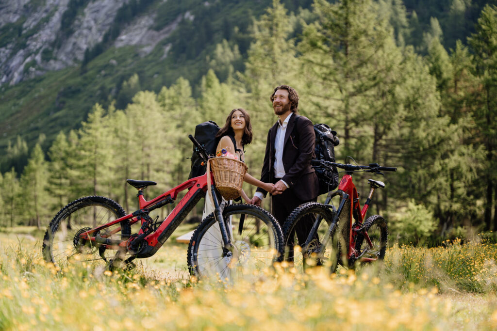A couple in white wedding dress and brown suit stand holding hands by two red mountain bikes, looking up at green valleys and tall Austrian mountains on their elopement day