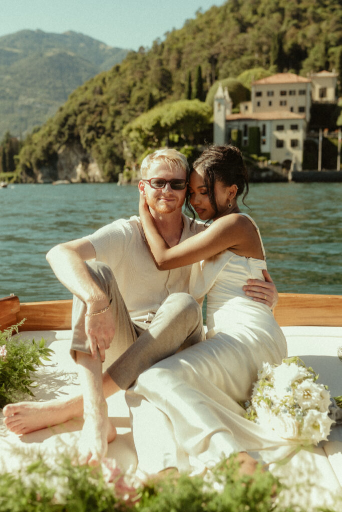 A mixed race couple cuddle on the back of a boat on Lake Como for their wedding photos, with Villa del Balbianello in the background