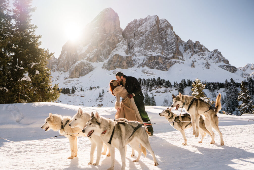 A couple kiss in front of a dog sled team with snowy mountain peaks behind them right after proposal