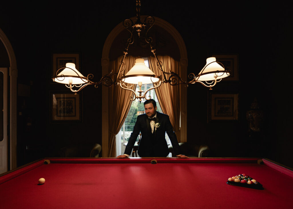 A man in blue suit leans over a red pool table in the Grand Hotel Tremezzo's pool room awaiting his partner on their Lake Como elopement day