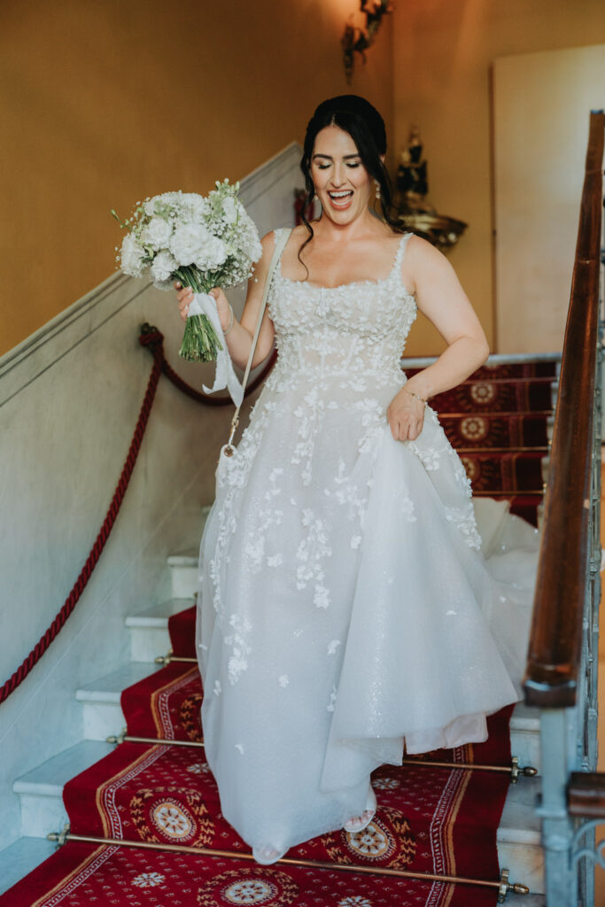A laughing bride holding her dress and a white bouquet descends red stairs at the Grand Hotel Tremezzo before her first look on her elopement day