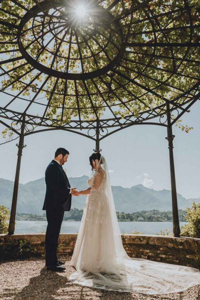 A couple in wedding attire stand under Villa Carlotta's rose gazebo exchanging rings on their Lake como Elopement day