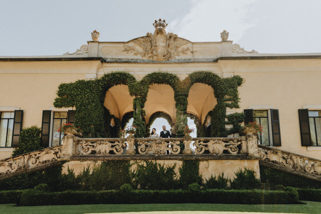 A couple in wedding attire stand on the balcony of the Villa del Balbianello Loggia surrounded by green arches 