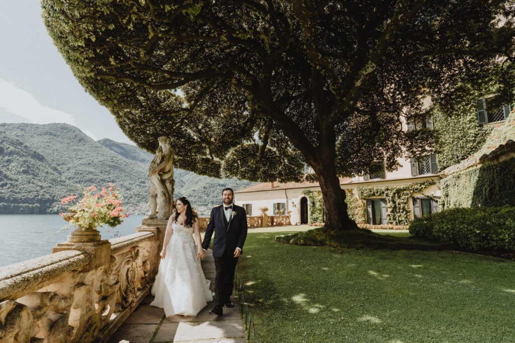 A couple in wedding attire walk far off on a pathway in front of Villa del Balbianello, Lake Como, for wedding photos
