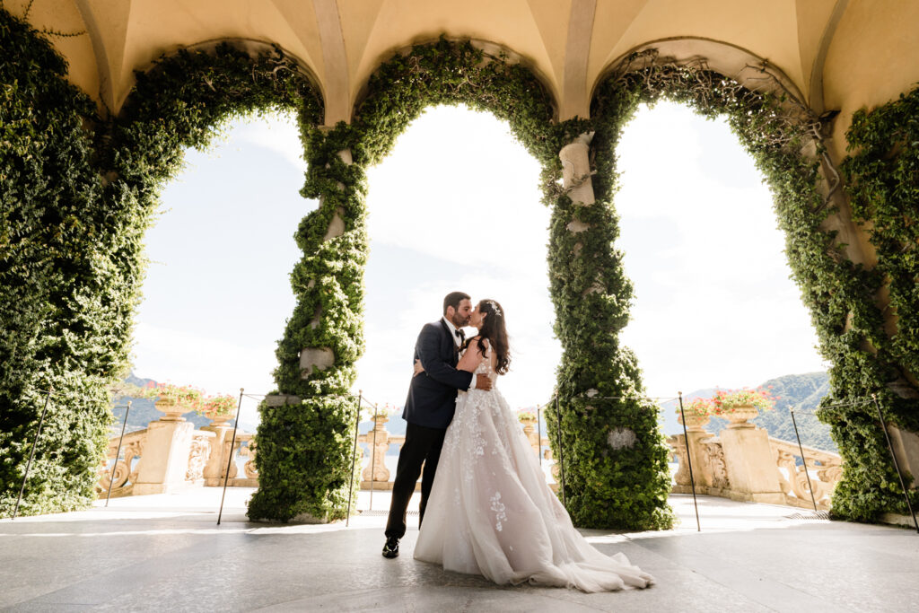 A couple in suit and white wedding dress kiss under the arches of the Loggia arches of Villa del Balbianello on their wedding day