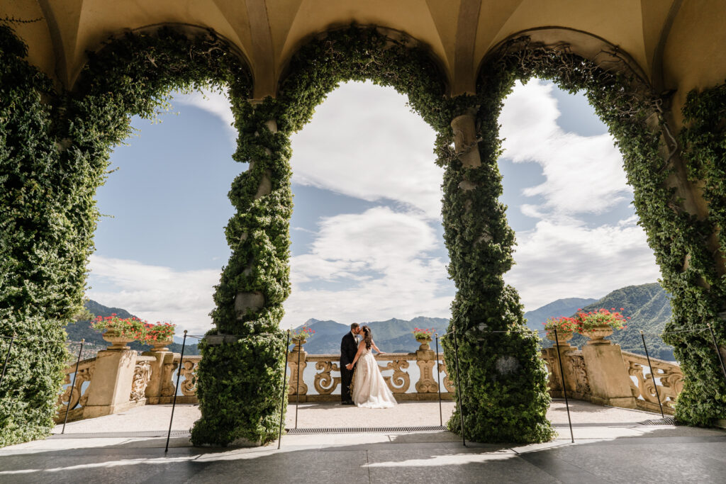 A couple in wedding dress and blue suit pose for their elopement photos in the arches of Villa del Balbianello, during their Lake Como elopement day