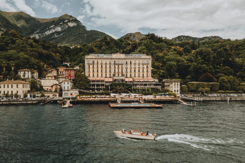 A couple in wedding attire sit on the back of a wooden boat on Lake Como with the Grand Hotel Tremezzo on the background during their elopement day