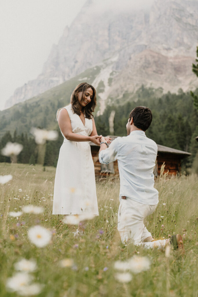 A man is one one knee proposing to his girlfriend in a wildflower meadow with colorful blooms and white daisies somewhere in the Dolomites