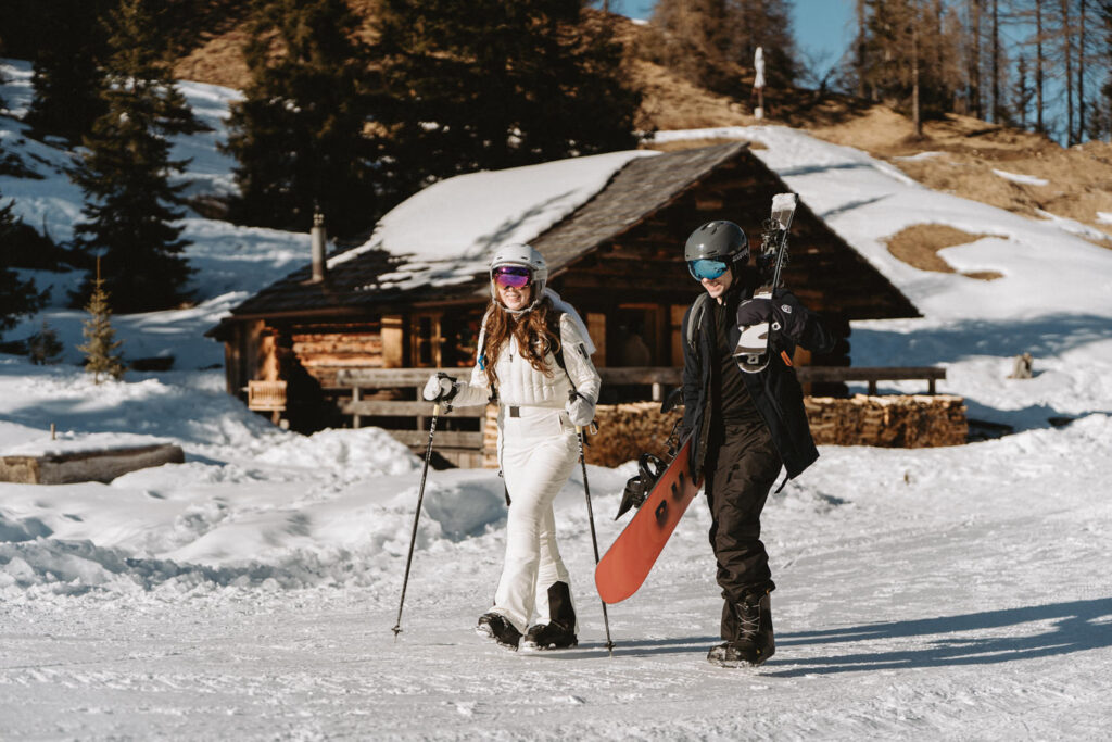A couple walk holding ski and snowboard gear before their winter proposal on the slopes