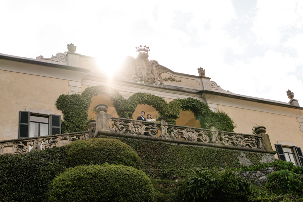 A couple stand under the Loggia arches of Villa del Balbianello, far in the distance, for their wedding photos