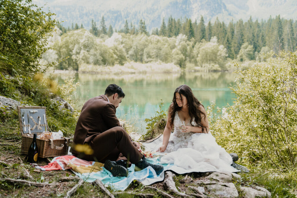 A couple in white wedding dress and brown suit sit near a turquoise lake in Slovenia, enjoying a picnic on their elopement day