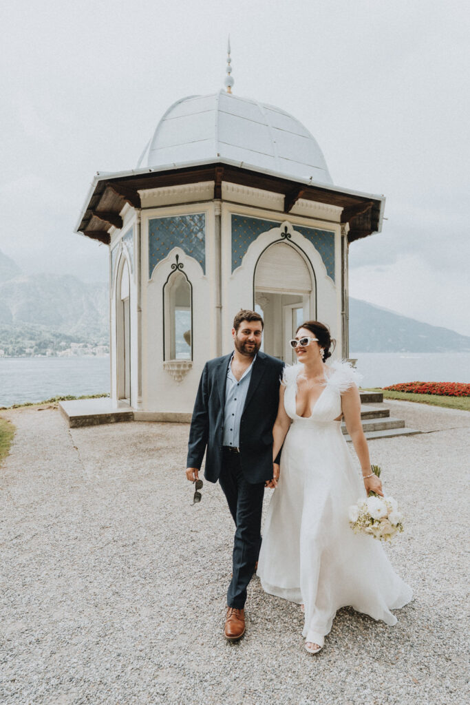 A couple in white wedding dress and blue suit walk casually toward the camera on the grounds of Villa Melzi, a lakeside villa on Lake Como popular for wedding and engagement photos